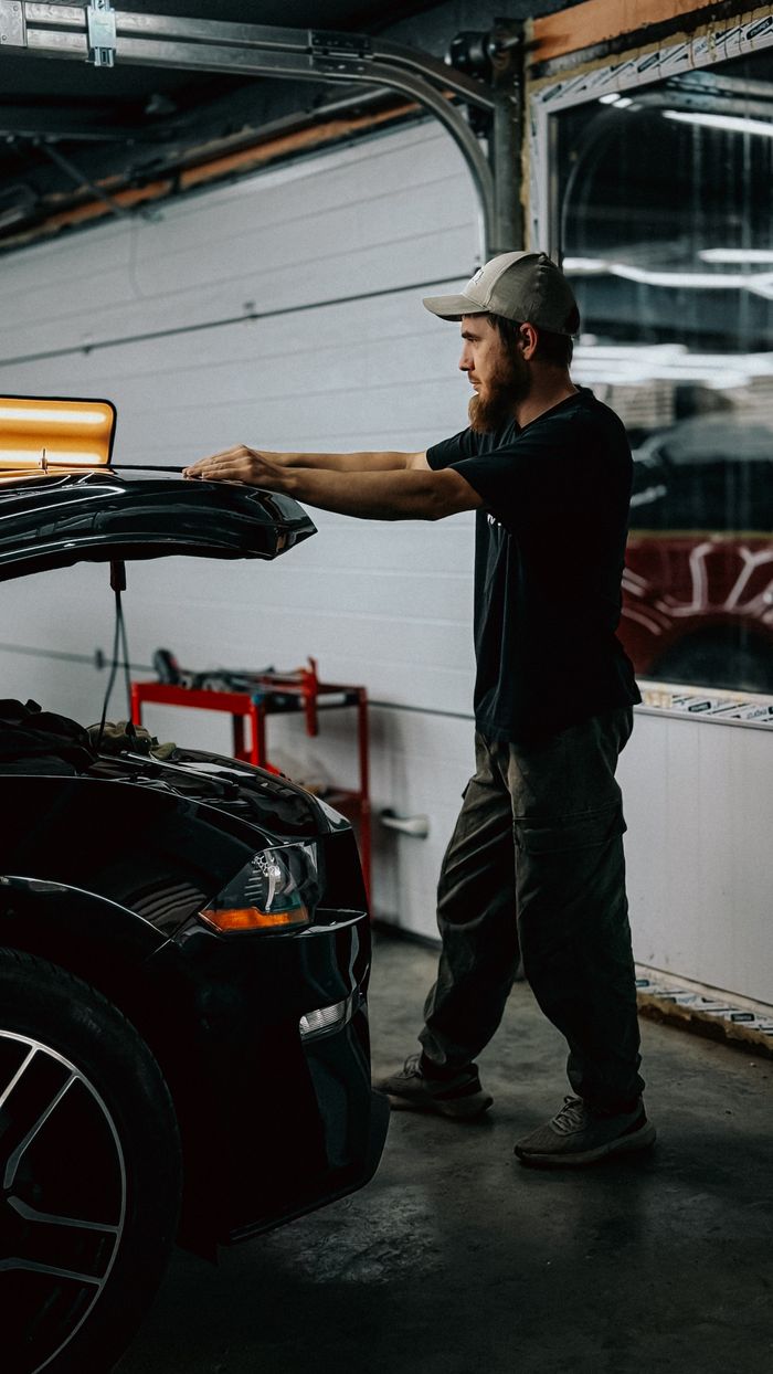 Young mechanic examines a car engine in a dimly lit garage setting, demonstrating auto repair work.
