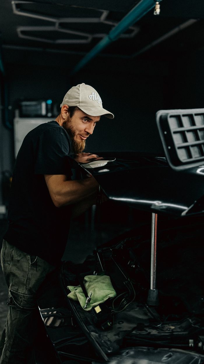 Young male mechanic works on a sleek car hood in a dimly lit garage.