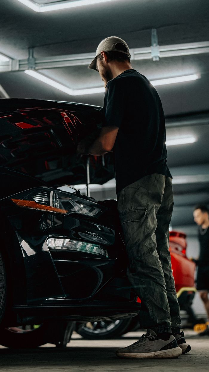 Mechanic inspecting car in a dimly lit garage with a focused expression, highlighting automotive repair and maintenance.